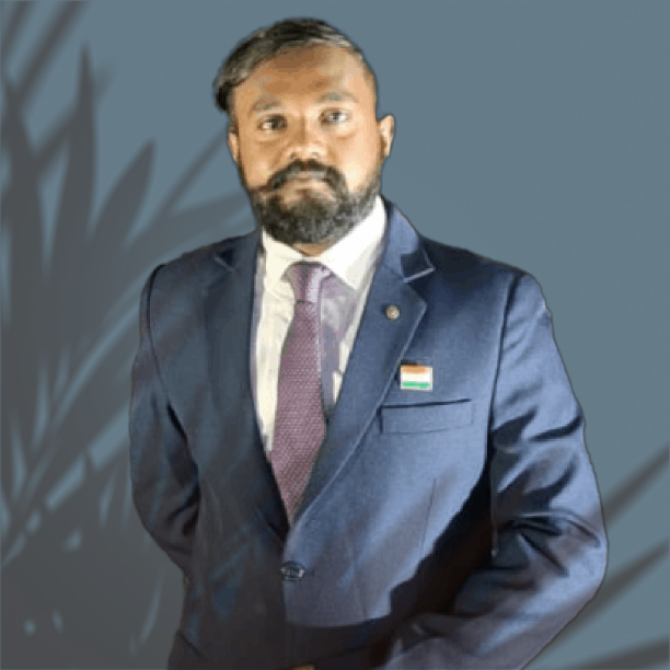 Mr. Dinesh Lal, CEO & Founder of Unicorniz Innovation. He is wearing a Blue Suit with Purple Tie on a White Shirt & a plant's shade falling on the background